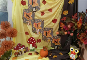 Dekoracja z okazji Powitania Jesieni - na tablicy pokrytej żółto-brązowym materiałem kolorowy napis "Jesień" oraz listki. Na szafce stoją sylwety sowy, jeża, muchomora oraz dynia. Obok szafki stoi wazon z kolorowymi, jesiennymi liśćmi.