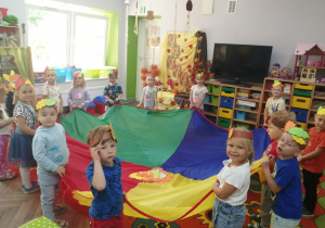 Dzieci ustawione w kole trzymają chustę animacyjną, na której znajdują się kolorowe listki. W tle dekoracja z okazji Powitania Jesieni.
