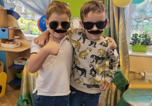 Kacper i Julek robią sobie pamiątkowe zdjęcie na tle dekoracji z okazji Dnia Chłopaka. Chłopcy mają na sobie doczepiane wąsy oraz okulary przeciwsłoneczne.