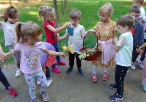 Grupa "Biedronek" na spacerze w parku podczas gromadzenia darów jesieni i obserwacji przyrodniczych. Franio i Ula trzymają kosz z kasztanami, żołędziami i jesiennymi listkami.