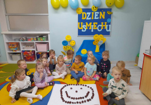 Przedszkolaki siedzą na dywanie, a przed nimi leży ułożona na kartonie uśmiechnięta buzia z kasztanów. W tle tablica z napisem Dzień Uśmiechu, a nad nią wiszą balony w kolorze żółtym i niebieskim.