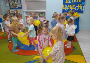 Dzieci tańczą przy muzyce w parach na dywanie. Przedszkolaki trzymają między sobą balony. W tke kącik konstrukcyjny, tablica z napisem Dzień Usmiechu.