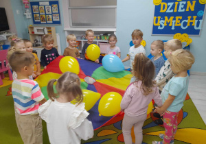 Dzieci stoją w kole i trzymają chustę animacyjną. Na chuście leżą balony w kolorze żółtym i niebieskim.