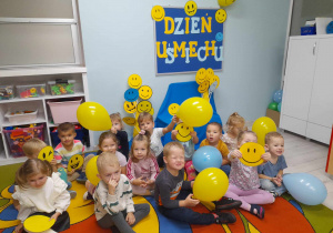 Dzieci siedzą na dywanie z balonami w kolorze żółtym i niebieskim oraz emblematami z wesołymi minkami. W tle tablica z napisem Dzień Uśmiechu.