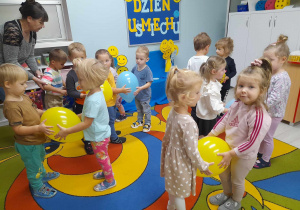 Dzieci w parach tańczą na dywanie trzymając między sobą balony w kolorze żółtym i niebieskim. Obok stoi Pani Grażynka. W tle dekoracja z okazji Dnia Uśmiechu.