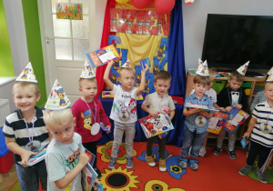 Grupka chłopców w papierowych czapeczkach na głowach stoją i trzymają upominki otrzymane przez Radę Rodziców: kolorowanki i kredki. W tle dekoracja.
