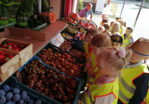 ”Motylki” stoją przed sklepem owocowo- warzywnym. W tle skrzynki z owocami i warzywami.