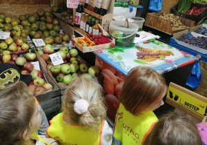 Grupka dzieci stoi w sklepie przed skrzynkami z jabłkami.