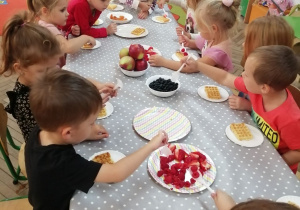 Grupka dzieci siedzi przy stoliku i dekoruje owocami gofry.
