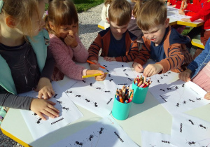 Julia, Natalia, Franek i Antek siedzą przy stolikach w kształcie plastra miodu i wypełniają karty pracy z wykorzystaniem kredek.