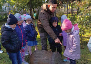 Dzieci razem z Panem leśnikiem stoją obok rzeźby żołędzia w ogrodzie przedszkolnym. W tle drzewa w jesiennych szatach.