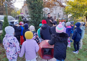 Dzieci stoją obok cisa w ogrodzie przedszkolnym, a Pan leśnik opowiada ciekawostki na temat rośliny. W tle budynek oraz drzewa w jesiennych szatach.
