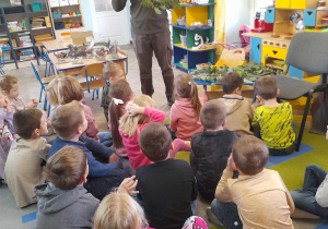Przedszkolaki siedzą na dywanie, a Pan leśnik stoi przed nimi i opowiada dzieciom o dębie prezentując jego gałązkę.