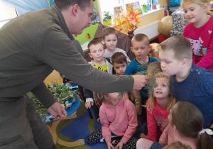 Dzieci siedzą na dywanie, a Pan leśnik daje im do powąchania gałązkę sosny.