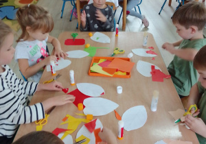 Dzieci z grupy Biedronek przy stolikach wycinają nożyczkami liście i wyklejają je papierem w jesiennych kolorach. Na stolikach: klej w sztyfcie, nożyczki, tacki z kolorowym papierem.