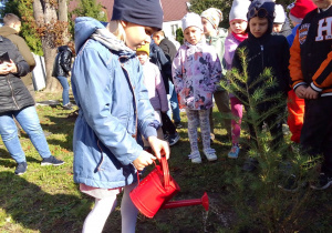 Ula czerwoną konewką podlewa zasadzone drzewko. Wokół dzieci z przedszkola obserwują działania Uli.