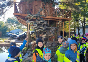 Dzieci z grupy Słoneczek idą ścieżką ekologiczną, za przedszkolakami widać domek na drzewie