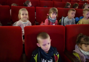Dzieci siedzą na czerwonych fotelach w sali projekcyjnej i oczekują na rozpoczęcie przedstawienia.