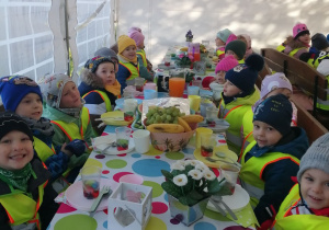 Zdjęcie grupowe dzieci w namiocie.Przedszkolaki siedzą przy wspólnym stole na którym znajdują się owocowe przekąski.
