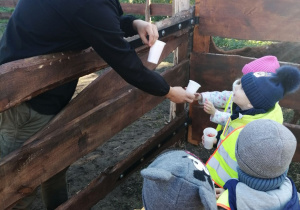 Grupka dzieci stoi przy ogrodzeniu i odbiera od pana kubeczki z marchewkami.