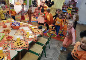 Dzieci podczas zabawy tanecznej. Tosia prowadzi „węża”. Obok stoją stoły na któtych znajdują się dyniowe serwetki, talerzyki oraz słodycze.