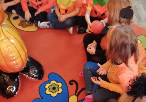 Dzieci siedzą w kole na dywanie i podają sobie małą dynię podczas zabawy "Dynia parzy".