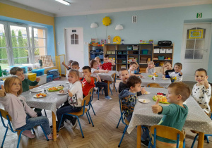 Dzieci siedzą przy stołach nakrytych ceratkami. Na środku każdego stołu leżą na talerzach owoce. Przedszkolaki podnoszą kciuki do góry. W tle kącik z układankami, okna, drzwi.