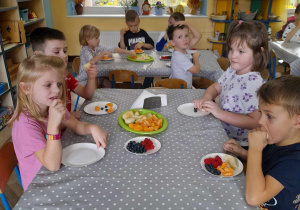 Dzieci przy dwóch stołach degustują owoce. W tle kącik przyrodniczy, okno.