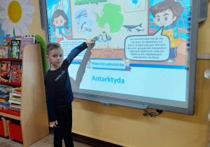 Oskar stoi przed tablicą multimedialną, na której wyświetlony jest slajd przedstawiający kontynent - Antarktydę. Chłopiec pokazuje na tablicy pingwiny.