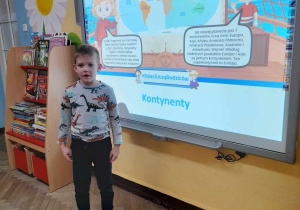 Filip z grupy "Słoneczka" stoi przed tablicą multimedialną, na której wyświetlony jest slajd przedstawiający kontynenty. Chłopiec wymienia nazwy kontynentów. Obok tablicy znajduje się kącik książki.