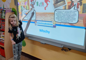 Lena stoi przed tablicą multimedialną, na której wyświetlony jest slajd przedstawiający Włochy. Dziewczynka pokazuje obrazek malarza.