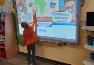 Chłopiec z grupy II "Motylki" stoi przed tablicą multimedialną, na której wyświetlony jest slajd przedstawiający kontynent - Afrykę. Chłopiec pokazuje na tablicy zwierzęta.