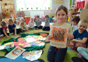 Dzieci w zgromadzone w kole wokół jesiennych liści i obrazków z ciekawostkami o jeżach. Jeden z obrazków prezentuje Lena.