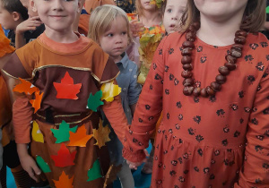 Dzieci w jesiennych strojach stoją w grupce obok siebie. Na pierwszym planie Oskar i Alicja trzymają się za ręce.