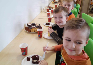 Dzieci siedzą przy stole podczas przerwy i jedzą ciasto na talerzykach.
