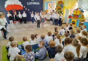 Dzieci ze wszystkich grup siedzą na dywanie i oglądają występ "Słoneczek".