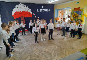 Dzieci z grupy "Słoneczka" stoją w półkolu w galowych strojach z kotylionami w barwach narodowych. Na środku sceny stoi Bartek, Alicja i Franek, którzy witają zaproszonych gości na uroczystości.