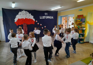 Dzieci tańczą Jesiennego Kujawiaka. Stoją w parach odwrócone do siebie po obwodzie koła, trzymają się za ręce i tupią nogami. W tle dekoracja z okazji Święta Niepodległości.