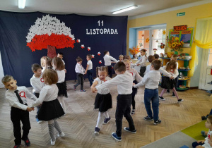 Dzieci w parach tańczą po obwodzie koła . W tle dekoracja z okazji Święta Niepodległości.