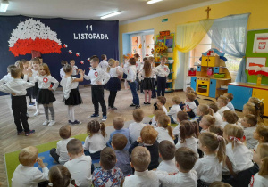 Dzieci ze wszystkich grup siedzą na dywanie, a przedszkolaki z grupy IV prezentują taniec na środku sceny. W tle dekoracja z okazji Święta Niepodległości.