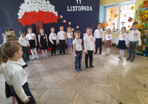 Dzieci stoją w półkolu podczas przedstawienia na tle dekoracji, a Eryk i Kacper na środku sceny mówią fragment wiersza.