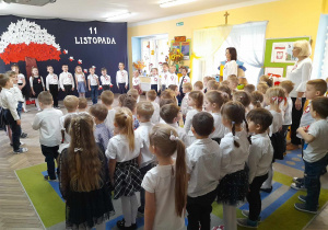 Dzieci z całego przedszkola, Pani dyrektor i Pani wicedyrektor śpiewają hymn narodowy. W tle dekoracja z okazji Święta Niepodległości.