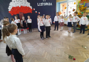 Dzieci stoją w półkolu na tle dekoracji, a na środku Anastazja i Gabriel deklamują wiersz. Przed dziećmi leża dwie róże - biała i czerwona.