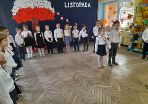 Dzieci strojach galowych stoją podczas występu na tle dekoracji. Wiktoria i Julek mówią fragment wiersza.