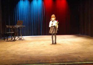 Zosia stoi na scenie w CTMiT i śpiewa piosenkę "Nasza Polska". Dziewczynka trzyma w prawej ręce mikrofon, a w lewej dwie róże - białą i czerwoną.
