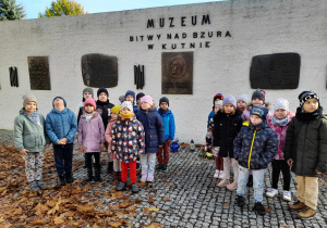 Dzieci stoją przed tablicami pamiątkowymi umieszczonymi na "Murze pamięci". Na murze znajduje się napis - Muzeum Bitwy nad Bzurą w Kutnie.