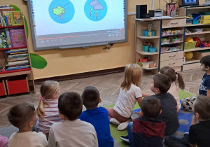 Dzieci siedzą na dywanie i rozwiązują zagadki słuchowe zaprezentowane na tablicy multimedialnej.