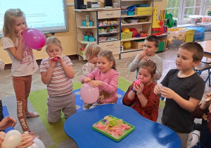 Dzieci nadmuchują balony, które się powiększają. Obok znajduje się stolik, na którym leżą piórka.