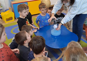 Dzieci zgromadzone są wokół stolika, na którym stoi szklane naczynie z wodą w kolorze niebieskim. Pani dyrektor umieszcza w nim mniejsze naczynie do góry dnem ze zgniecioną kartką papieru.