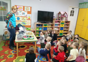 Dzieci siedzą na dywanie, a przed nimi stoi przy stole Pani Magdalena. Pani prezentuje przedszkolakom maszynę do szycia i opowiada o zawodzie krawcowej.
